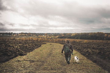 The hunter is walking across the field, with weapons. Next to the dog is a springer spaniel. Autumn