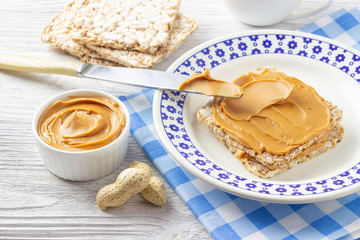 Creamy peanut butter. Paste in bowl, sandwich and peanuts on white wooden table.