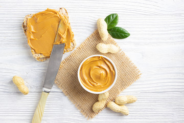 Creamy peanut butter. Paste in bowl, sandwich and peanuts on white wooden background. Above view.