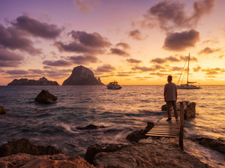 Fototapeta premium atardecer de otoño en la playa y embarcadero de Cala D´hort con vistas a Es Vedra. Hombre observando los barcos y el paisaje