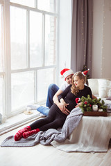 An attractive pregnant woman and a handsome man in a Santa hat embrace on a white bed