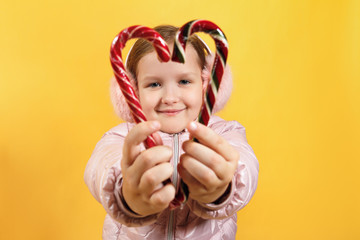 Happy little girl holding heart shaped christmas candy. A child in a jacket and warm ear muffs on a yellow background. The concept of Christmas, New Year