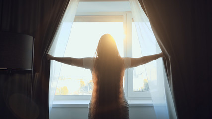 Young woman opens dark curtains and enjoys morning sunlight sun, view from back