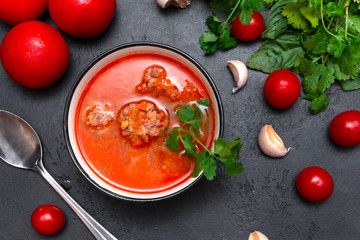 black plate of red tomato soup with meat meatballs, cilantro on a black background, spoon, cloves and garlic head, fresh cherry , top view,