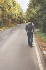 A young man with a backpack behind him is walking along an empty autumn road. Travel, hipster. Walk in the fall, autumn trees.