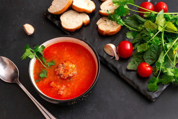 1 black plate of red tomato soup with meat meatballs, greens on a black background, spoon, garlic, fresh cherry tomatoes, top view