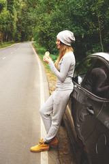 A young woman is eating a burger near a car on an empty road. Food on the trip. Food on the go. Autumn travel. Fast food.