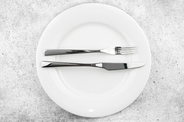 Table setting. Empty plate, knife and fork on a light concrete background. The fork and knife lie on a plate in parallel, the meal is finished, I liked the dish. Top view and flat lay with copy space.