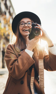 Smiling girl in hat travels in holiday. Photographer in glasses with retro photo camera. Tourist portrait. Sunlight street in europe city. Traveler hipster shooting architecture, copy space mockup