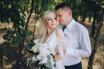 Wedding portrait of lovers newlyweds on the background of nature and the forest with trees. Stylish groom hugs a beautiful blonde bride in a white dress with a bouquet. Concept and photography.