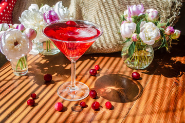 Still life with glass of red drink with berries and ice surrounded by white flowers on a textile background on a wooden table in the diagonal shadow of the blinds on a bright sunny day. Greeting card