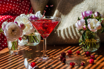 Still life with glass of red drink with berries with a splash and splatters of falling berry surrounded by flowers on textile background in striped shadow. Beautiful light refraction on table setting