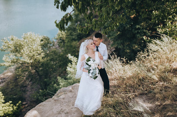 Wedding portrait of beautiful newlyweds on a background of greenery, rocks and a river in nature. Stylish groom hugs a cute blonde bride in a white dress. Photography and concept.