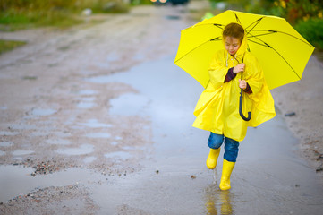 Obraz premium Cute little girl in a raincoat and with a yellow umbrella in her hands walks through puddles, walks in boots and squirts
