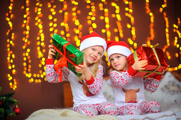 Two cute little girls in pajamas and santa claus hats are sitting on the bed with gifts in their hands. Cristmas presents.