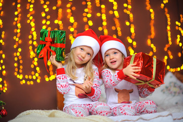 Two cute little sisters in pajamas are sitting on the bed with gifts in their hands. Cristmas presents.