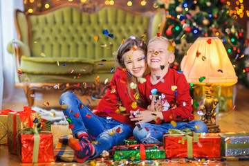 Happy children are sitting on the floor near gifts under the falling candy and hugging, against the background of a Christmas tree and holiday lights. Cristmas presents.