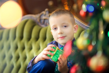 Christmas time, cute little boy with a gift sits on a sofa near the festive Christmas tree, smiling happy children