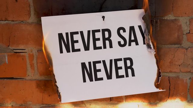The inscription on a white A4 size sheet against the background of an orange brick wall. The paper with the black word Never Say Never burns, smoke and turns into ashes. Business concept.