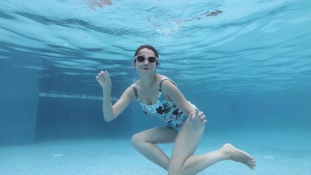 Cute middle aged woman swimming and posing underwater in pool in different poses on Sunny day. She looks at the camera in sunglasses and smiles at the bottom of the pool. Slow motion. 4K.