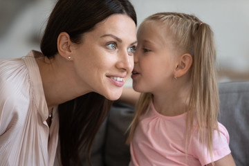 Little daughter telling secret to smiling mother, whispering in ear