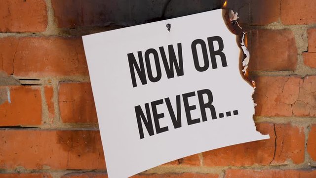 The inscription on a white A4 size sheet against the background of an orange brick wall. The paper with the black word Now or Never burns, smoke and turns into ashes. Business concept.