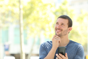 Happy adult pensive man holding phone looks at side