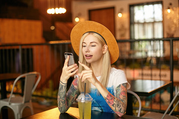 Young beautiful woman with blonde long hair posing over modern cafe interior in white and blue t-shirt and wide hat, keeping smartphone in hands and checking social networks