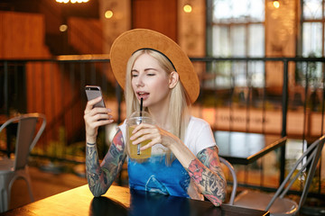Portrait of pretty young long haired female with tattooes sitting at table in cafe with modern interior and drinking lemonade, holding smartphone in raised hand and looking at screen