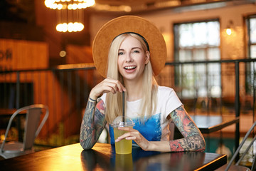 Cheerful blonde long haired woman with tattooes keeping hands on table while drinking fresh juice with straw, looking at camera positively and smiling joyfully