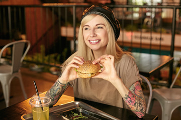 Horizontal shot of pretty tattooed blonde female in beige t-shirt and leather black beret posing over modern cafe interior, looking aside with wide charming smile and holding burger in hands