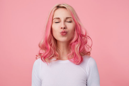 Portrait of lovely positive female with pink curly hair posing over pink background and smiling sincerely, keeping eyes closed and folding lips in kiss, wearing casual white t-shirt