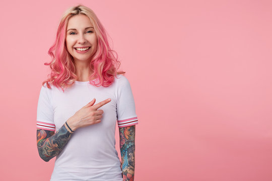 Joyful tattooed pretty lady with pink curly hair wearing casual clothes, standing over pink background with raised hand and pointing aside with forefinger, smiling joyfully to camera