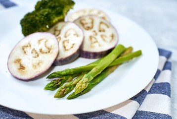 Healthy eating concept. Steamed vegetables in a white plate on a blue table. Eggplant, broccoli, asparagus.