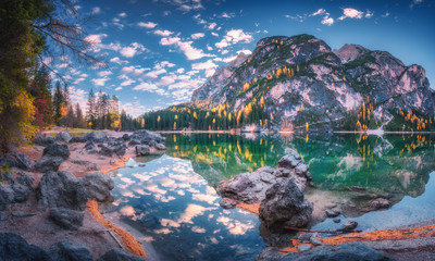 Beautiful Braies lake at sunrise in autumn in Dolomites, Italy. Landscape with mountains, blue sky with clouds, water with reflection, stones, trees with colorful leaves. Lake in fall. Panorama - obrazy, fototapety, plakaty