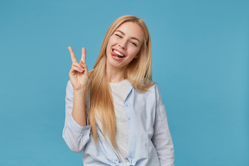 Joyful young blonde woman with long hair fooling over blue background in casual clothes, showing tongue cheerfully to camera and raising two fingers with victory gesture