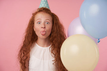 Close-up of funny redhead girl with curly hair in white dress and birthday cap celebrates something, joyfully looking in camera and showing tongue. Posing over pink background with colored ballons