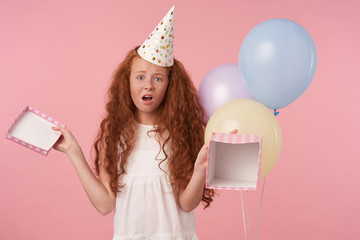 Portrait of cute girl with foxy curly hair in elegant dress and birthday cap celebrates holiday, being disappointed to get empty birthday present, isolated over pink studio background