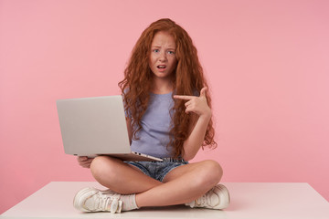 Obraz na płótnie Canvas Unpleased pretty female teenager with foxy curly hair raising hand and showing aside with forefinger, sitting with crossed legs over pink background and holding modern laptop