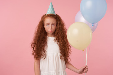 Horizontal shot of upset redhead little girl with long curly hair wearing festive clothes and birthday cap, looking at camera sadly and pursing lips, isolated over pink background with air balloons