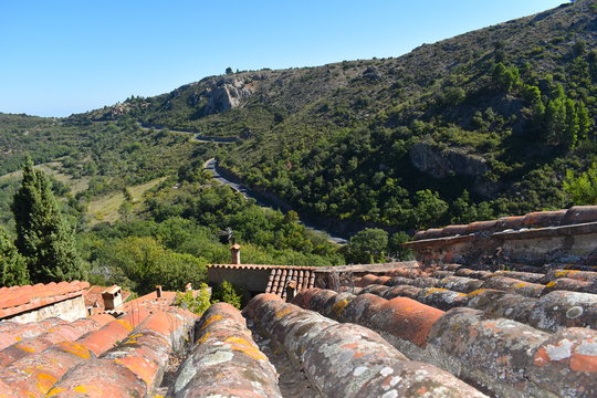 Brown, red, orange and yellow French roof tiles and chimneys overlooking winding mountain road surrounded by greenery. Picturesque self drive European family holiday. 