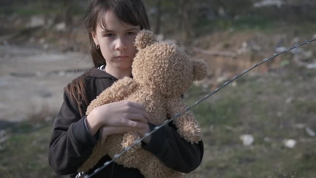 Alone with toy. A homeless little girl hugs a bear cub in a refugee camp. Childhood without parents.