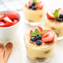 homemade, exquisite dessert tiramisu in glasses decorated with strawberry, blueberry, mint on white wooden table