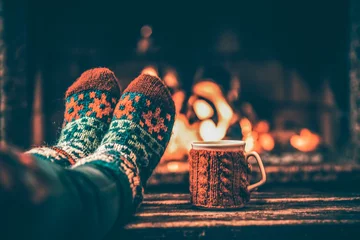Foto auf Leinwand Füße in Wollsocken am Weihnachtskamin. Frau entspannt sich bei warmem Feuer mit einer Tasse heißem Getränk und wärmt ihre Füße in Wollsocken auf. Auf den Füßen hautnah. Winter- und Weihnachtsferienkonzept. © AlexMaster