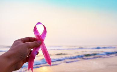 Breast cancer awareness month concept, hands holding pink ribbon against beach with blue sky. Blur...