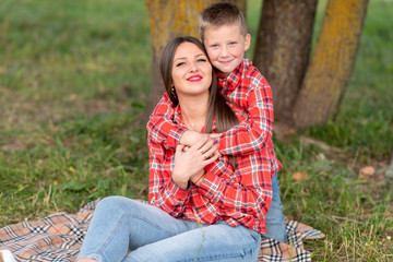 The son gently hugs his mother's shoulders, both smiling, sitting on a checkered bedspread.