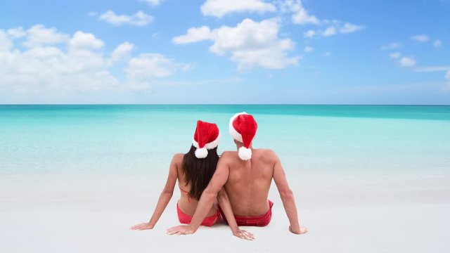CINEMAGRAPH - seamless loop: Christmas couple in love lying down relaxing on white sand beach sun tanning in tropical travel destination in winter holidays. Back view of young adults wearing santa hat