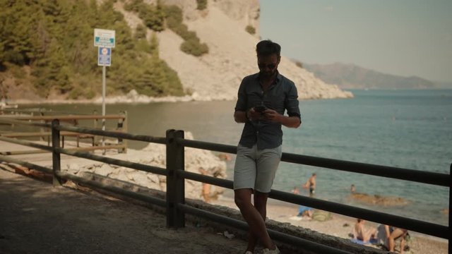 Male Chatting With Friends.Man Using Mobile Phone On Vacation In Sunny Day.Man Having Chat On Smartphone At Mediterranean Beach.Relaxed Man Looking And Texting On Mobile Phone In Vocation Near Sea.