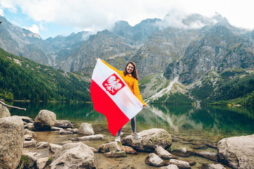 young pretty smiling woman holding poland flag at beach of mountain lake