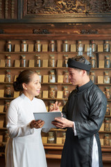 Pharmacist of old Asian drugstore showing infomation on tablet computer to coworker and giving him tasks for the day
